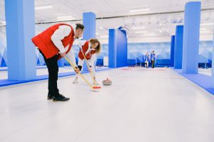 Two players in red vests sweeping in front of a curling stone on an indoor ice rink.
