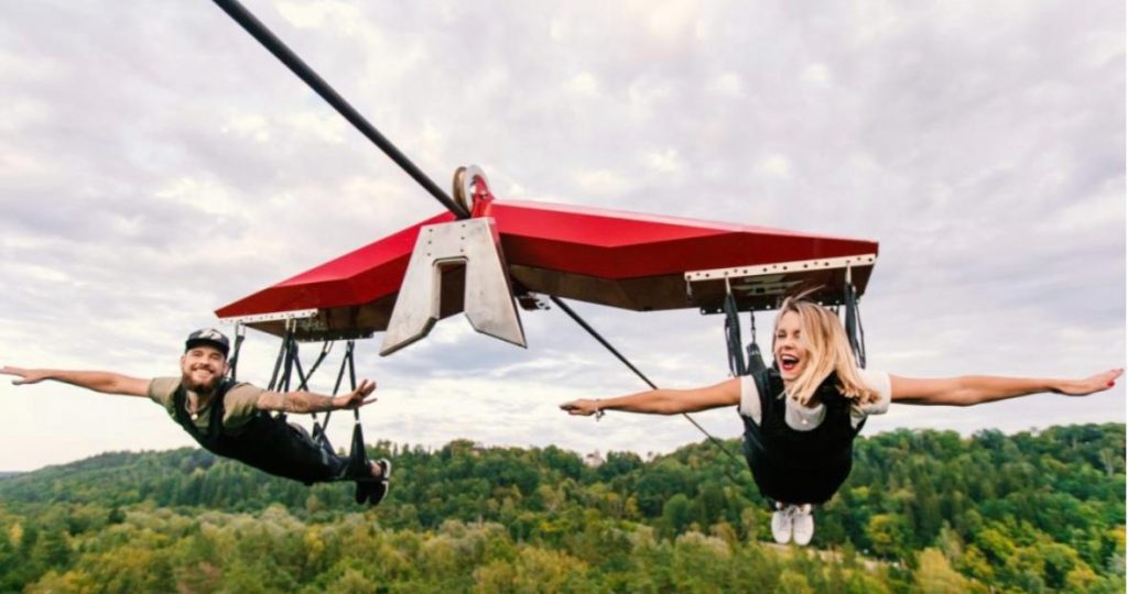 Two people flying on a red zipline glider over a forest.