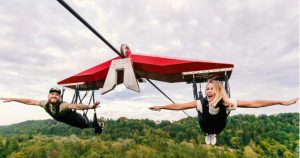 Two people flying on a red zipline glider over a forest.