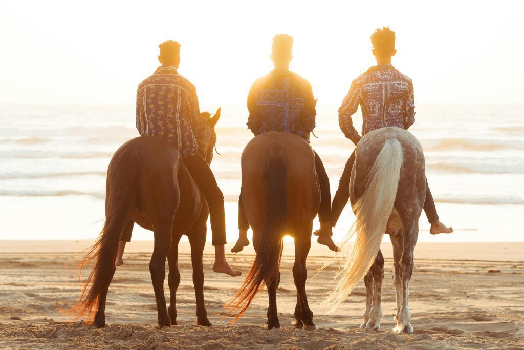 Three men on horseback facing the ocean at sunset.