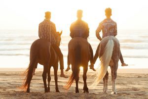 Three men on horseback facing the ocean at sunset.